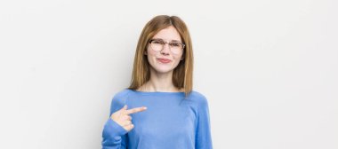 Kızıl saçlı güzel bir kadın gururlu, kendinden emin ve mutlu görünüyor, gülümsüyor ve kendini işaret ediyor ya da bir numara işareti yapıyor.