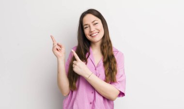 pretty caucasian woman smiling happily and pointing to side and upwards with both hands showing object in copy space