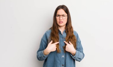 pretty caucasian woman pointing to self with a confused and quizzical look, shocked and surprised to be chosen