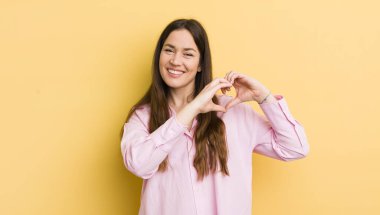 pretty caucasian woman smiling and feeling happy, cute, romantic and in love, making heart shape with both hands