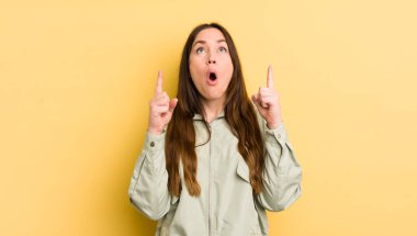 pretty caucasian woman looking shocked, amazed and open mouthed, pointing upwards with both hands to copy space