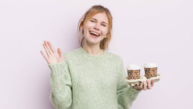 Kızıl saçlı güzel kız mutlu bir şekilde gülümsüyor, el sallıyor, seni selamlıyor ve selamlıyor. kahve konseptini kaldır.