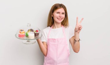 Kızıl saçlı güzel kız gülümsüyor ve arkadaş canlısı görünüyor. İki numarayı gösteriyor. Ev yapımı kek konsepti