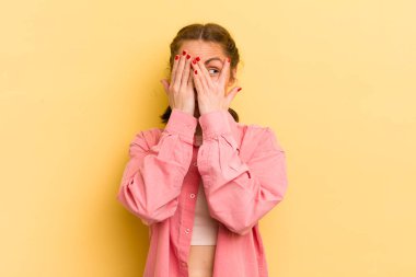 Genç ve güzel bir kadın elleriyle yüzünü kapatıyor, parmaklarının arasından şaşkın bir ifadeyle yan tarafa bakıyor.