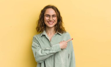 Genç, güzel bir kadın neşeli bir şekilde gülümsüyor, mutlu hissediyor ve kenarı ve yukarıyı işaret ediyor, kopyalama alanında nesneler gösteriyor.