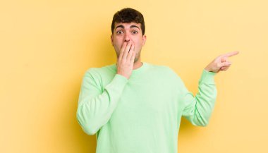 Genç yakışıklı adam kendini mutlu, şaşkın ve şaşırmış hissediyor. Ağzını eliyle kapatıyor ve yanal kopya alanını işaret ediyor.