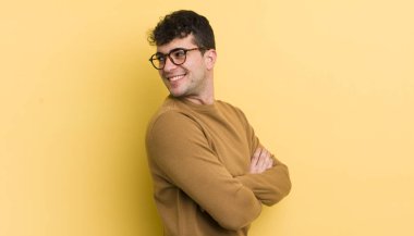 Genç yakışıklı adam neşeyle gülümsüyor, mutlu, tatmin olmuş ve rahatlamış hissediyor, kollarını kavuşturup yan tarafa bakıyor.