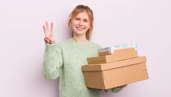赤頭の女の子は笑顔で友好的に見える3番目を示している 配送ボックスのコンセプト — ストック写真
