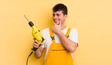 Genç ve yakışıklı bir adam mutlu ve kendinden emin bir ifadeyle gülümsüyor, elleri çenesinde. Tamirci ve matkap konsepti