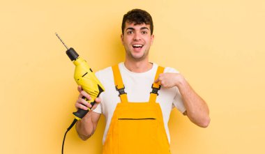 Genç ve yakışıklı bir adam mutlu hissediyor ve heyecanla kendini işaret ediyor. Tamirci ve matkap konsepti