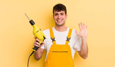 Genç yakışıklı adam mutlu bir şekilde gülümsüyor, el sallıyor, sizi selamlıyor ve selamlıyor. Tamirci ve matkap konsepti