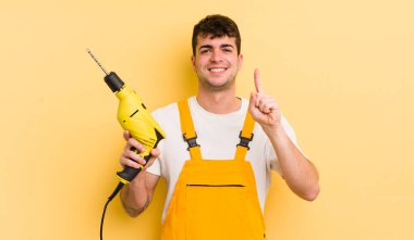 Genç yakışıklı adam gülümsüyor ve arkadaş canlısı görünüyor, bir numarayı gösteriyor. Tamirci ve matkap konsepti