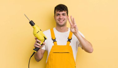 Genç yakışıklı adam gülümsüyor ve arkadaş canlısı görünüyor, üç numarayı gösteriyor. Tamirci ve matkap konsepti