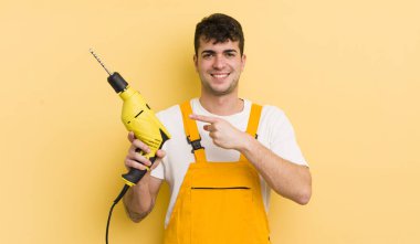 Genç yakışıklı adam neşeyle gülümsüyor, mutlu hissediyor ve bir tarafı işaret ediyor. Tamirci ve matkap konsepti