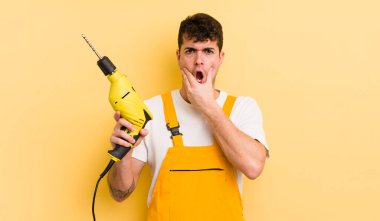 Ağzı ve gözleri açık ve eli çenesinde genç yakışıklı bir adam. Tamirci ve matkap konsepti