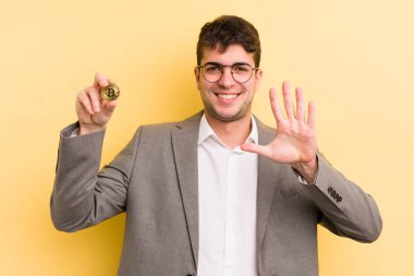 Genç yakışıklı adam gülümsüyor ve arkadaş canlısı görünüyor, beş numarayı gösteriyor. Bitcoin konsepti