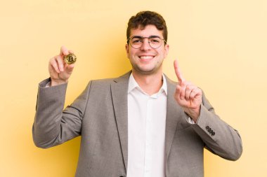 Genç yakışıklı adam gülümsüyor ve arkadaş canlısı görünüyor, bir numarayı gösteriyor. Bitcoin konsepti