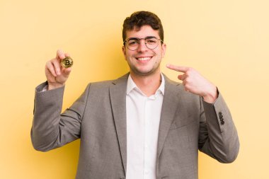 Genç ve yakışıklı bir adam kendi gülüşünü işaret ederek kendine güvenerek gülümsüyor. Bitcoin konsepti