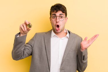 Genç, yakışıklı adam şaşırmış ve şok olmuş görünüyor. Çenesi düşmüş, elinde bir nesne var. Bitcoin konsepti