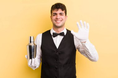 Genç yakışıklı adam mutlu bir şekilde gülümsüyor, el sallıyor, sizi selamlıyor ve selamlıyor. barmen kokteyli