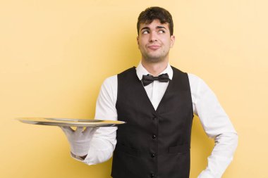 Genç yakışıklı adam omuz silkiyor, kafası karışık ve kararsız. garson ve tepsi konsepti