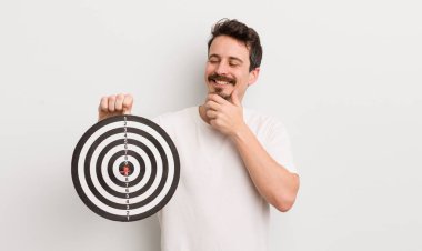 Genç ve yakışıklı bir adam mutlu ve kendinden emin bir ifadeyle gülümsüyor, elleri çenesinde. dart hedef konsepti