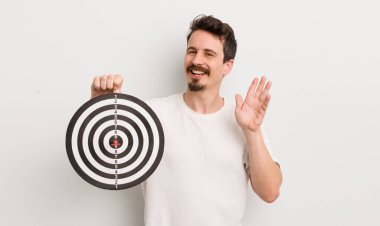Genç yakışıklı adam mutlu bir şekilde gülümsüyor, el sallıyor, sizi selamlıyor ve selamlıyor. dart hedef konsepti