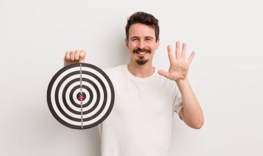 Genç yakışıklı adam gülümsüyor ve arkadaş canlısı görünüyor, beş numarayı gösteriyor. dart hedef konsepti