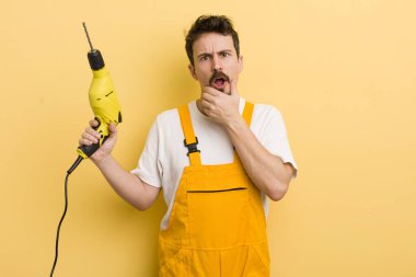 Ağzı ve gözleri açık ve elleri çenesinde. Matkap konsepti olan tamirci.