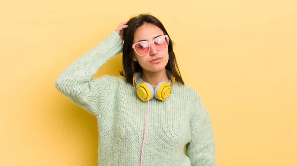 若いヒスパニック系の女性は困惑し 混乱し 頭を引っ掻いて感じている ヘッドフォンとサングラスのコンセプト — ストック写真