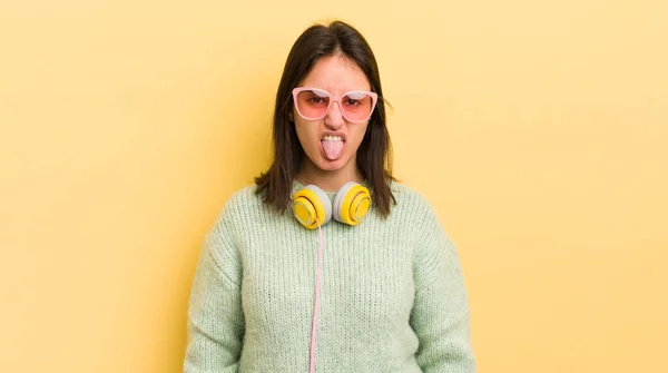 若いヒスパニック系の女性はうんざりしていらいらして舌を出す感じ ヘッドフォンとサングラスのコンセプト — ストック写真
