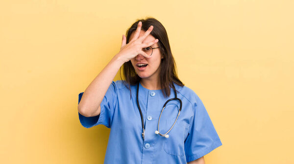 young hispanic woman looking shocked, scared or terrified, covering face with hand. nurse concept