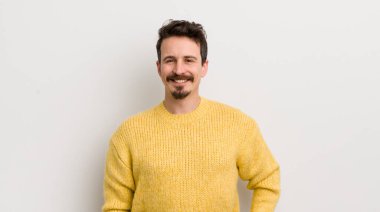 İspanyol genç adam mutlu bir şekilde gülümsüyor, eli kalçasında ve kendinden emin, olumlu, gururlu ve arkadaş canlısı bir tavırla.