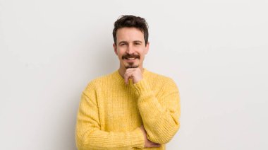 İspanyol asıllı genç adam mutlu ve gülümsüyor, elleri çenesinde, merak ediyor ya da soru soruyor, seçenekleri karşılaştırıyor.