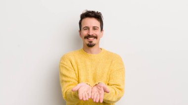 İspanyol genç adam mutlu bir şekilde gülümsüyor, arkadaş canlısı, kendinden emin, olumlu bakışlı, bir nesne ya da konsept sunuyor ve gösteriyor.