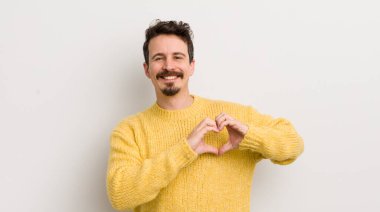 İspanyol genç adam gülümsüyor ve mutlu, sevimli, romantik ve aşık, iki eliyle kalbi şekillendiriyor.