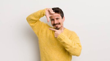 İspanyol genç adam mutlu, arkadaş canlısı ve pozitif hissediyor, gülümsüyor ve elleriyle bir portre veya fotoğraf çerçevesi yapıyor.