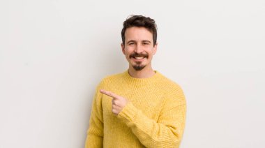 İspanyol genç adam neşeyle gülümsüyor, mutlu hissediyor ve kenarı ve yukarıyı işaret ediyor, fotokopi alanında nesneler gösteriyor.