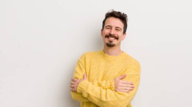 İspanyol genç adam kollarını kavuşturup mutlu bir şekilde gülüyor, rahat, pozitif ve tatmin edici bir pozla.