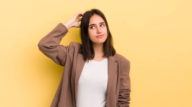 Genç İspanyol kadın şaşkın ve şaşkın hissediyor, kafasını kaşıyor ve yan tarafa bakıyor.