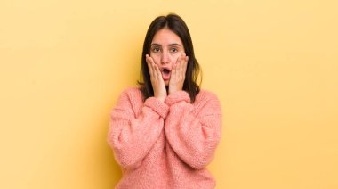 Genç İspanyol kadın şok olmuş ve korkmuş hissediyor, ağzı açık ve elleri yanaklarında korkmuş görünüyor.