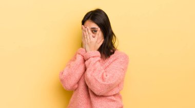 Latin kökenli genç bir kadın korkmuş ya da utanmış hissediyor. Gözleri yarı kapalı ya da elleri kapalı gözetliyor.