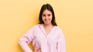 Genç İspanyol kadın mutlu bir şekilde gülümsüyor, eli kalçasında ve kendinden emin, olumlu, gururlu ve arkadaş canlısı bir tavırla.