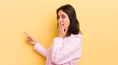 Genç İspanyol kadın mutlu, şaşkın ve şaşırmış hissediyor. Ağzını eliyle kapatıyor ve yanal fotokopi alanını işaret ediyor.