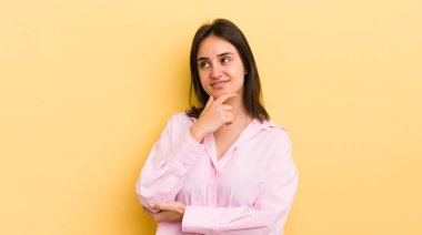 Genç İspanyol kadın mutlu, kendinden emin bir ifadeyle gülümsüyor, çenesinde elleri, merak ediyor ve yan tarafa bakıyor.