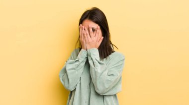 Latin kökenli genç bir kadın korkmuş ya da utanmış hissediyor. Gözleri yarı kapalı ya da elleri kapalı gözetliyor.