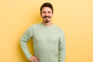 Genç İspanyol adam mutlu bir şekilde gülümsüyor, eli kalçasında ve kendinden emin, olumlu, gururlu ve arkadaş canlısı bir tavırla.