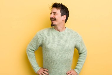 Genç İspanyol adam mutlu, neşeli ve kendinden emin görünüyor. Gururla gülümsüyor ve iki elini de kalçasında istiyor.