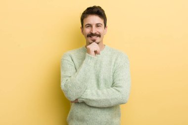 Genç İspanyol adam mutlu görünüyor ve gülümsüyor. Elleri çenesinde, merak ediyor ya da soru soruyor. Seçenekleri karşılaştırıyor.