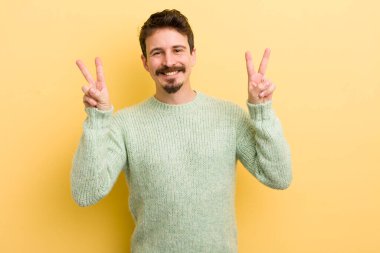 Genç İspanyol adam gülümsüyor ve mutlu görünüyor, arkadaş canlısı ve tatmin olmuş, iki eliyle zafer veya barış işareti yapıyor.